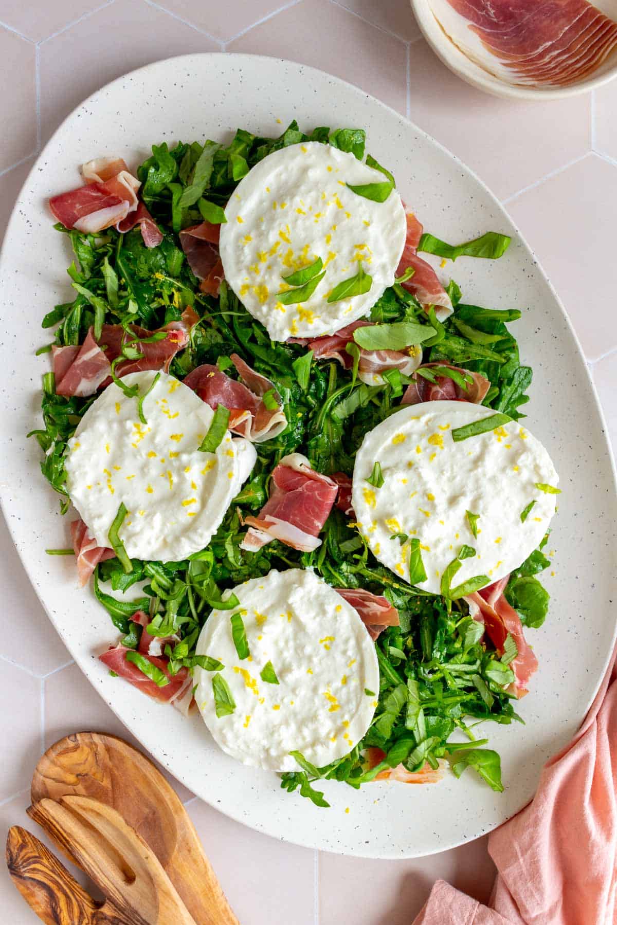Overhead view of a platter with arugula, prosciutto, burrata, and basil.