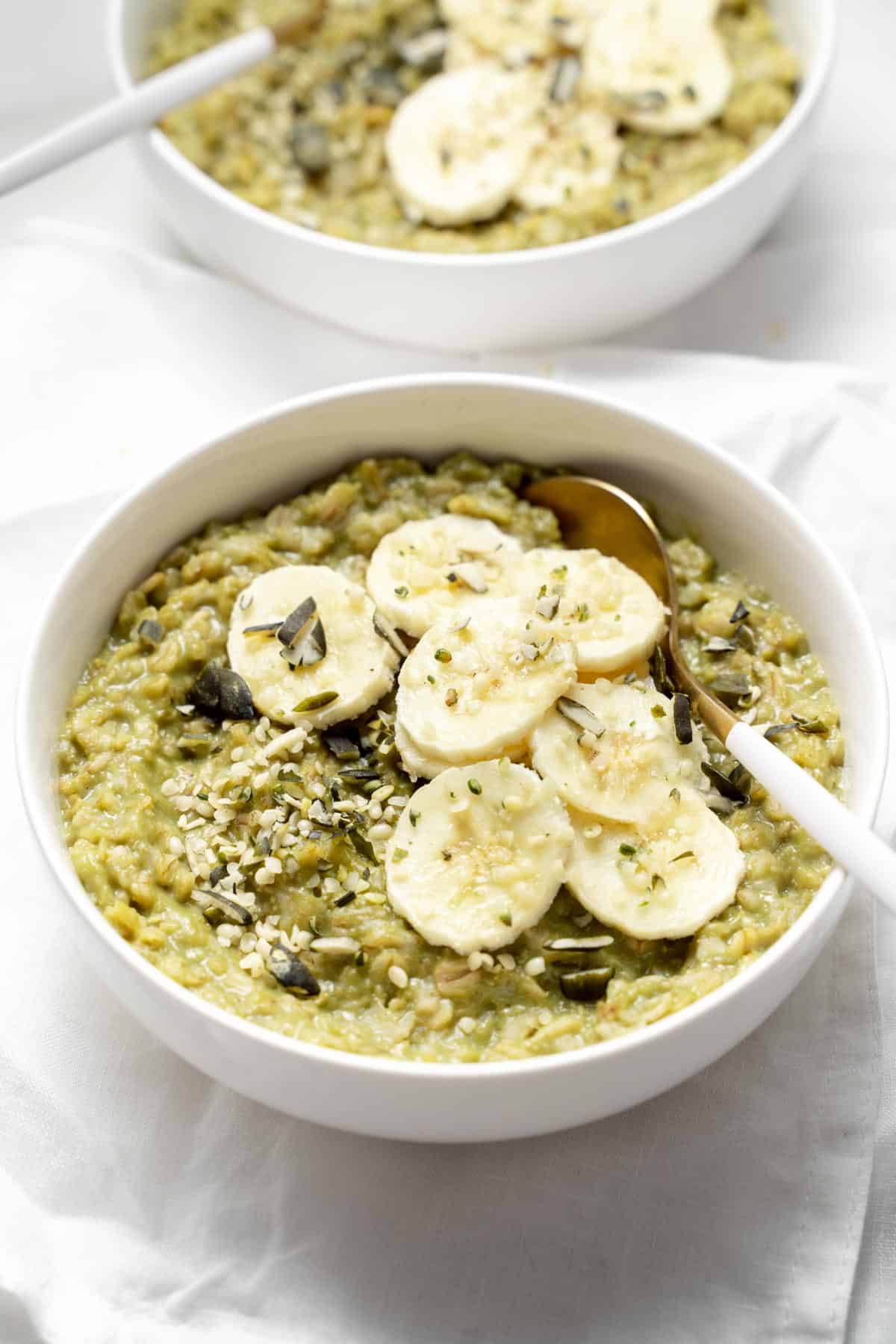 An angled view of a bowl of matcha oatmeal with sliced bananas and hemp seeds on top with a spoon tucked in.