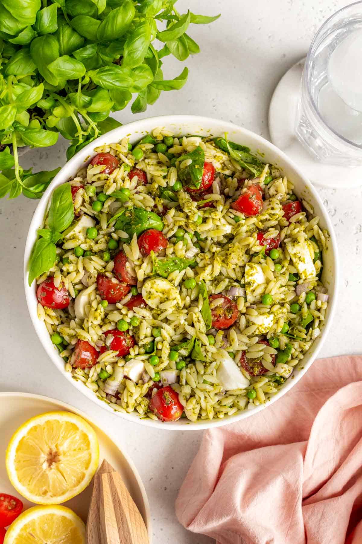 Overhead view of a large bowl of pesto orzo salad with some basil, water, and lemons around it.