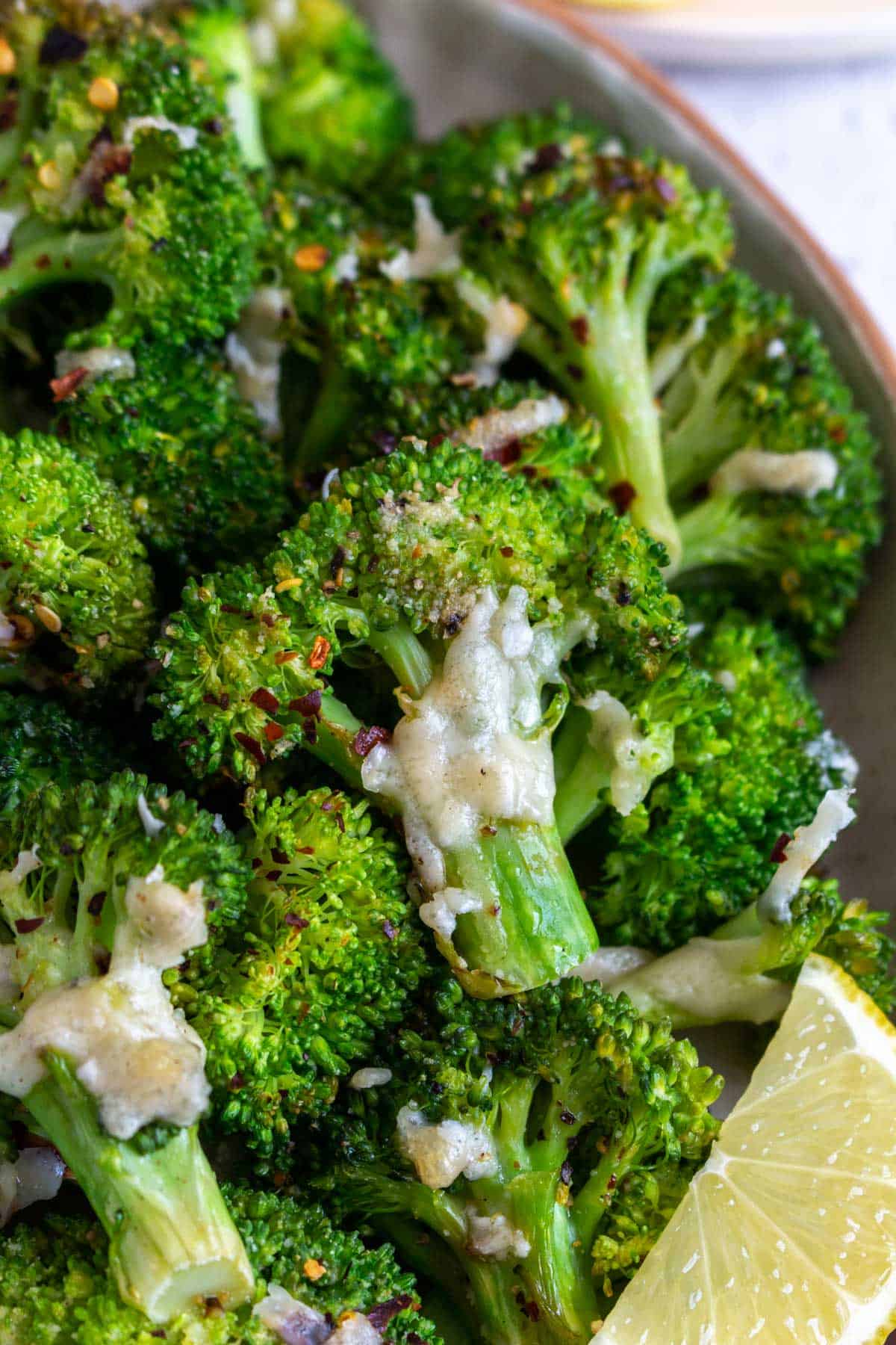 A close up view of smashed broccoli with red pepper flakes and melted parmesan and a lemon wedge.