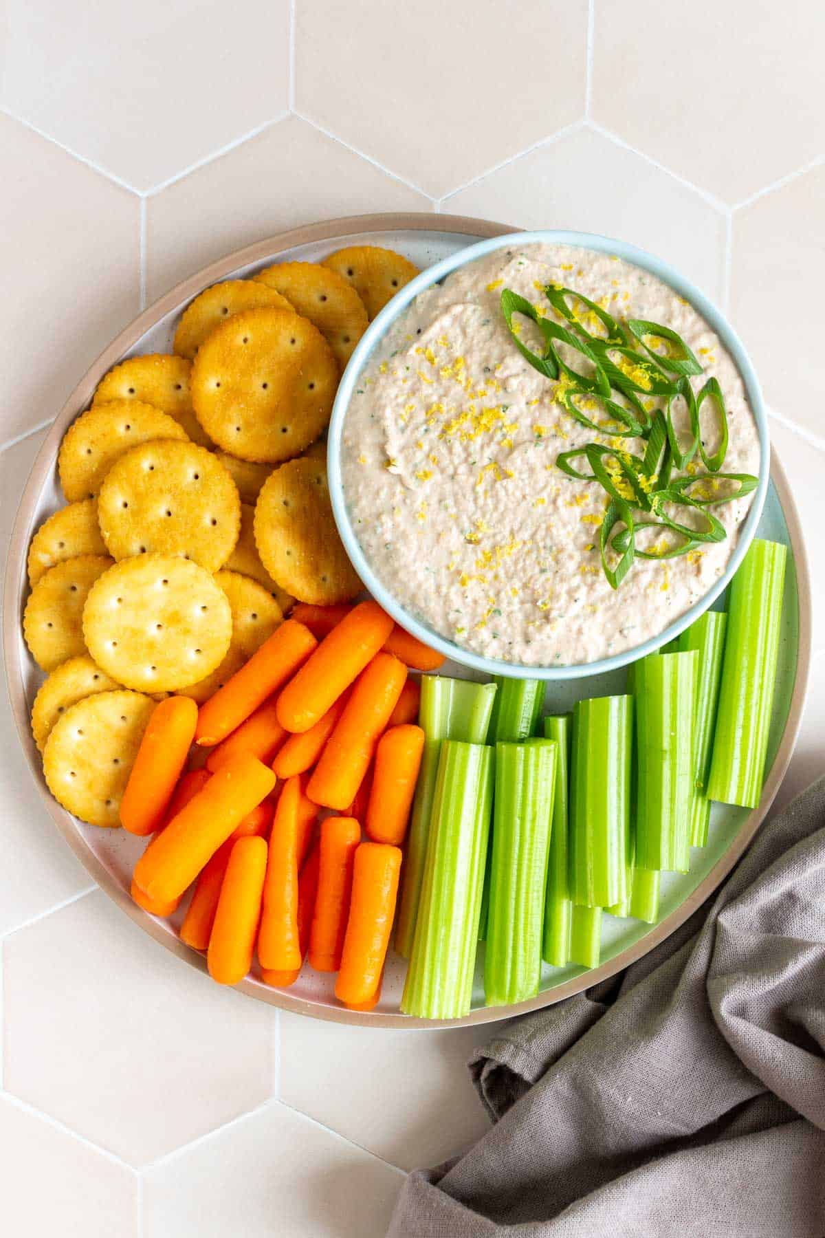 A plate of crackers, carrots, and celery stick with a bowl of tuna dip.