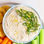 A bowl of tuna dip topped with lemon zest and green onions with some celery sticks, baby carrots, and crackers beside it.