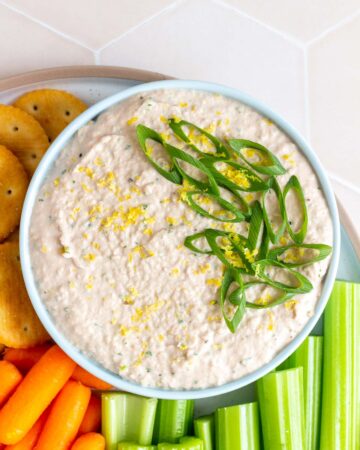 A bowl of tuna dip topped with lemon zest and green onions with some celery sticks, baby carrots, and crackers beside it.
