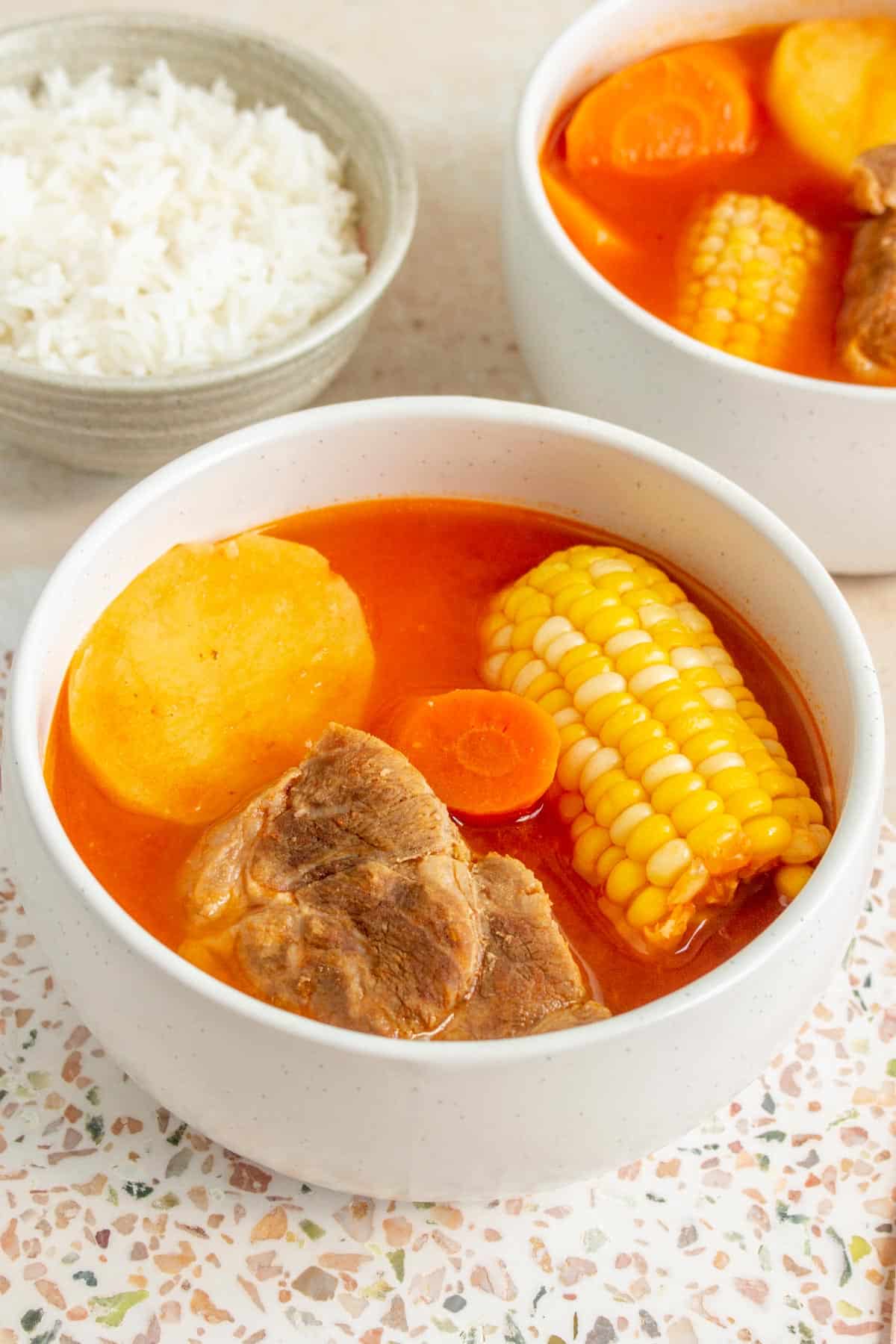 A bowl of soup with pork, corn on the cob, carrot, and potato with a bowl of rice in the background and another bowl of soup.