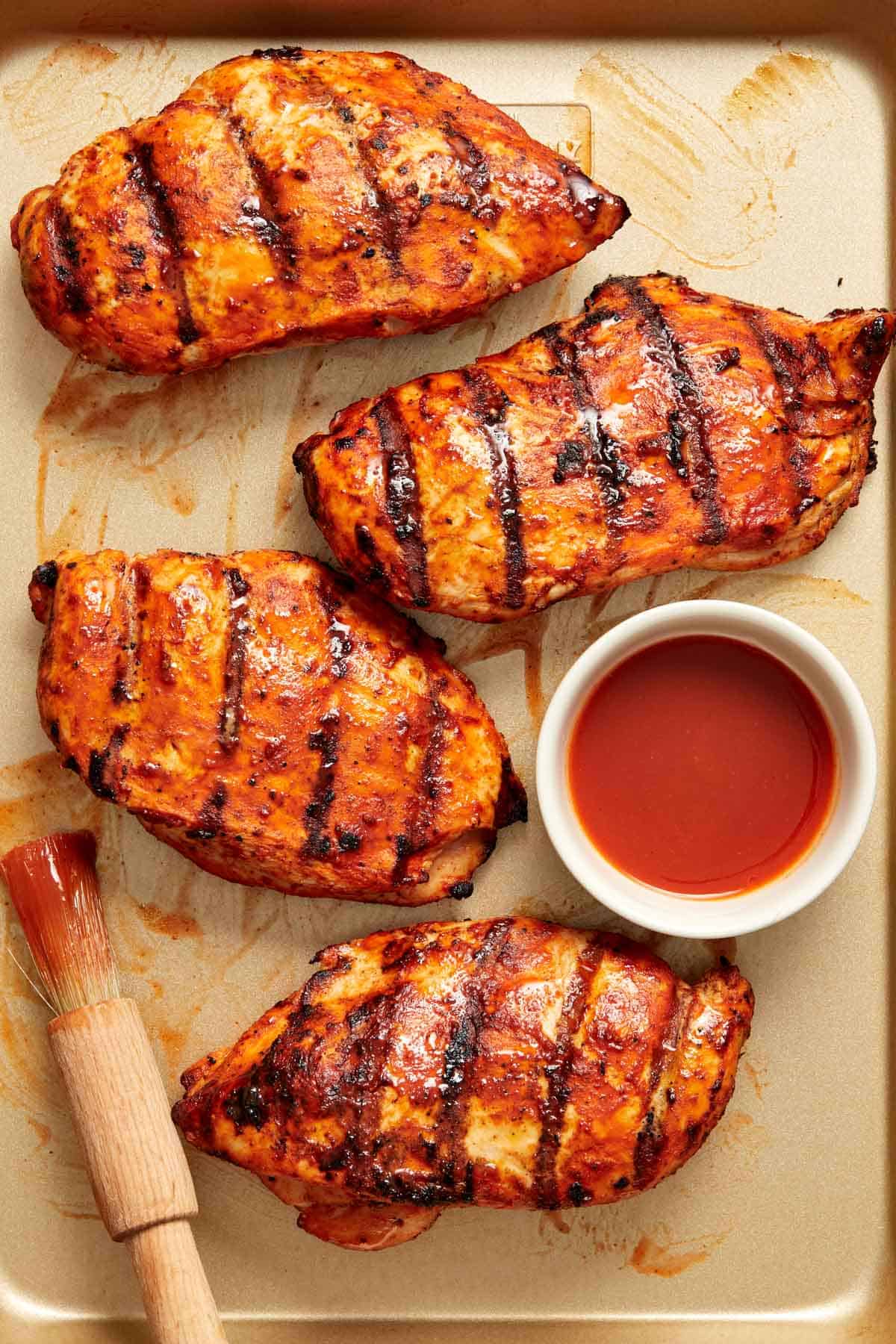 Overhead view of four grilled buffalo chicken breasts with a bowl of buffalo sauce and brush beside them.