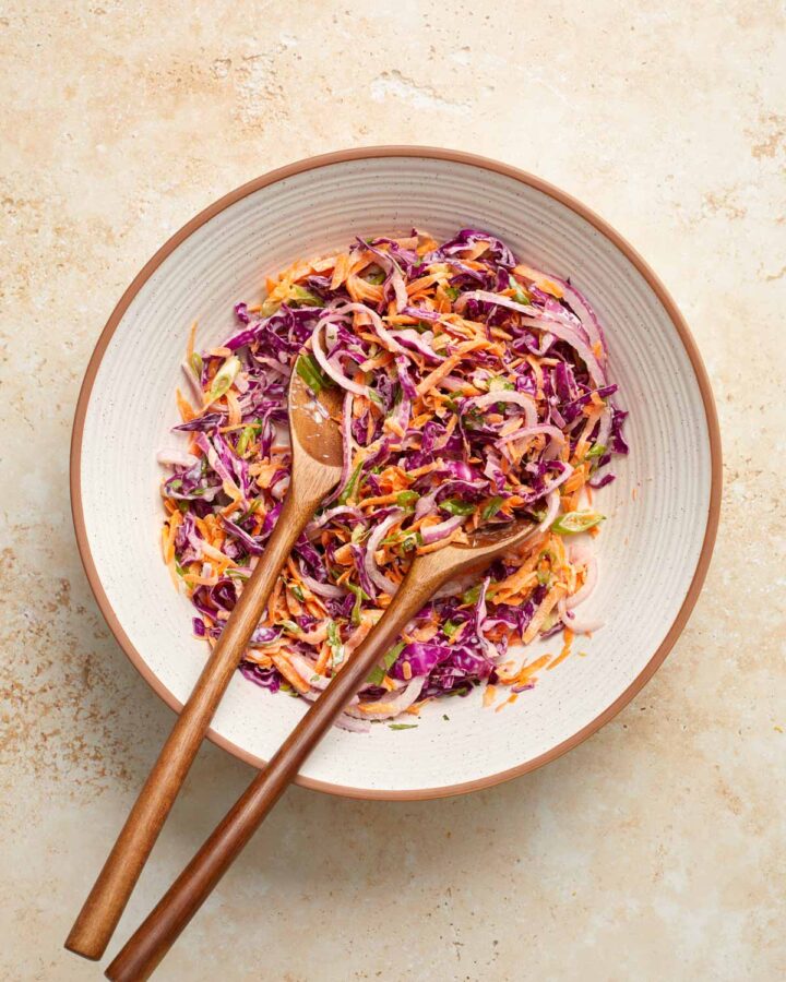 Overhead view of a bowl of purple cabbage slaw with wooden serving spoons inside.