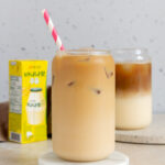 A glass with iced banana latte on a coaster with a banana milk box in the back.