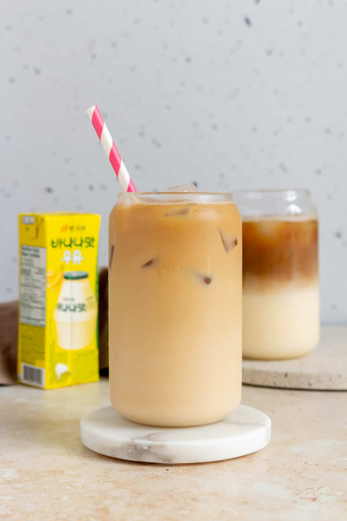 A glass with iced banana latte on a coaster with a banana milk box in the back.