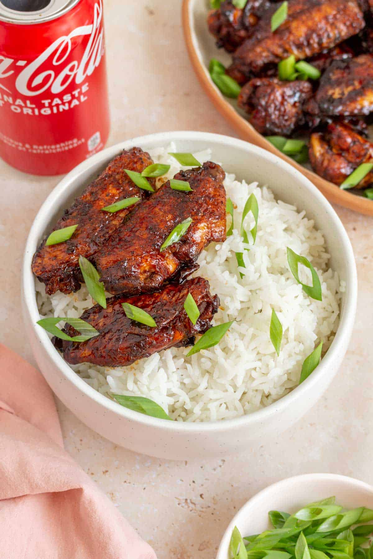 A bowl of rice with three cola chicken wings with green onions with a can of cola in the background.