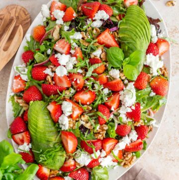 Overhead view of a platter of strawberry goat cheese salad. Fresh basil, bowl of goat cheese, scattered pecans and strawberries beside the platter.