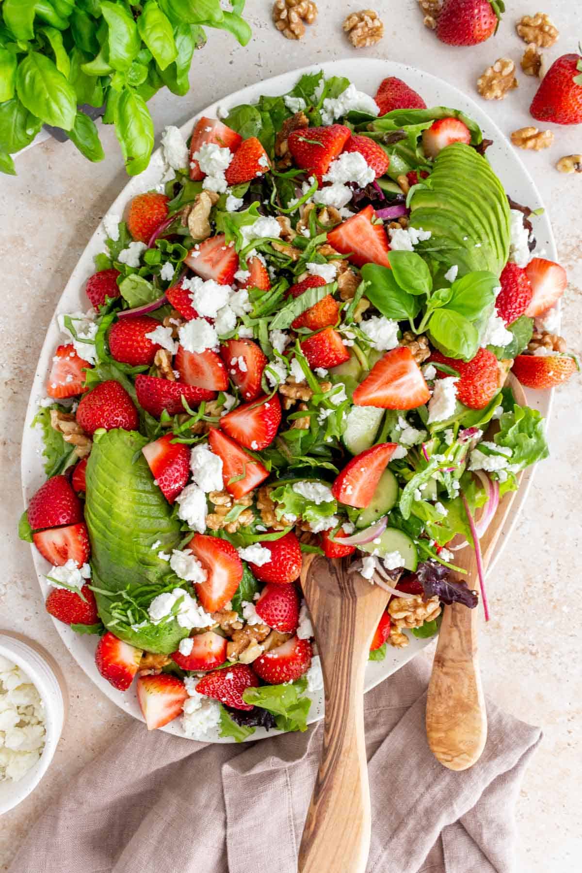 Overhead view of a large serving platter of strawberry goat cheese salad with avcado and wooden serving spoons inside the salad.
