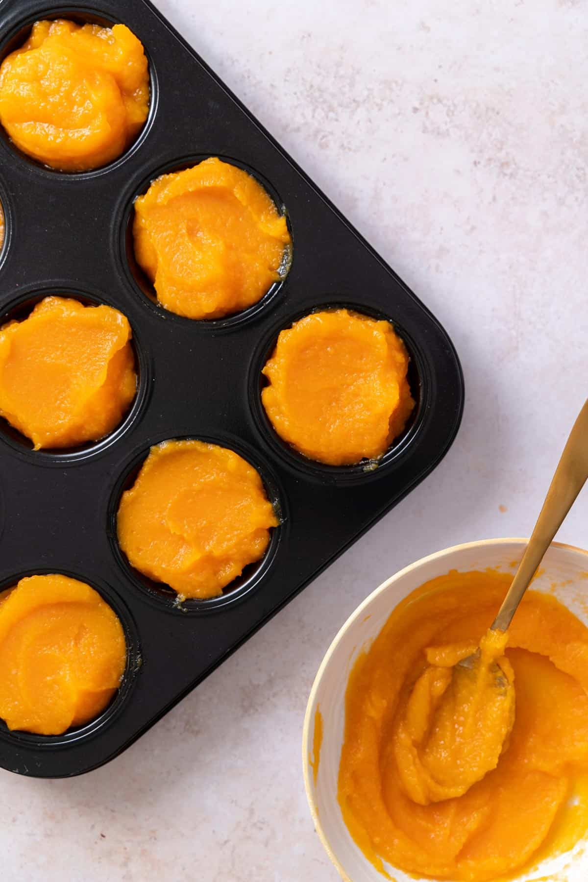 Muffin tin filled with homemade pumpkin puree with a bowl of more puree beside it with a spoon inside.