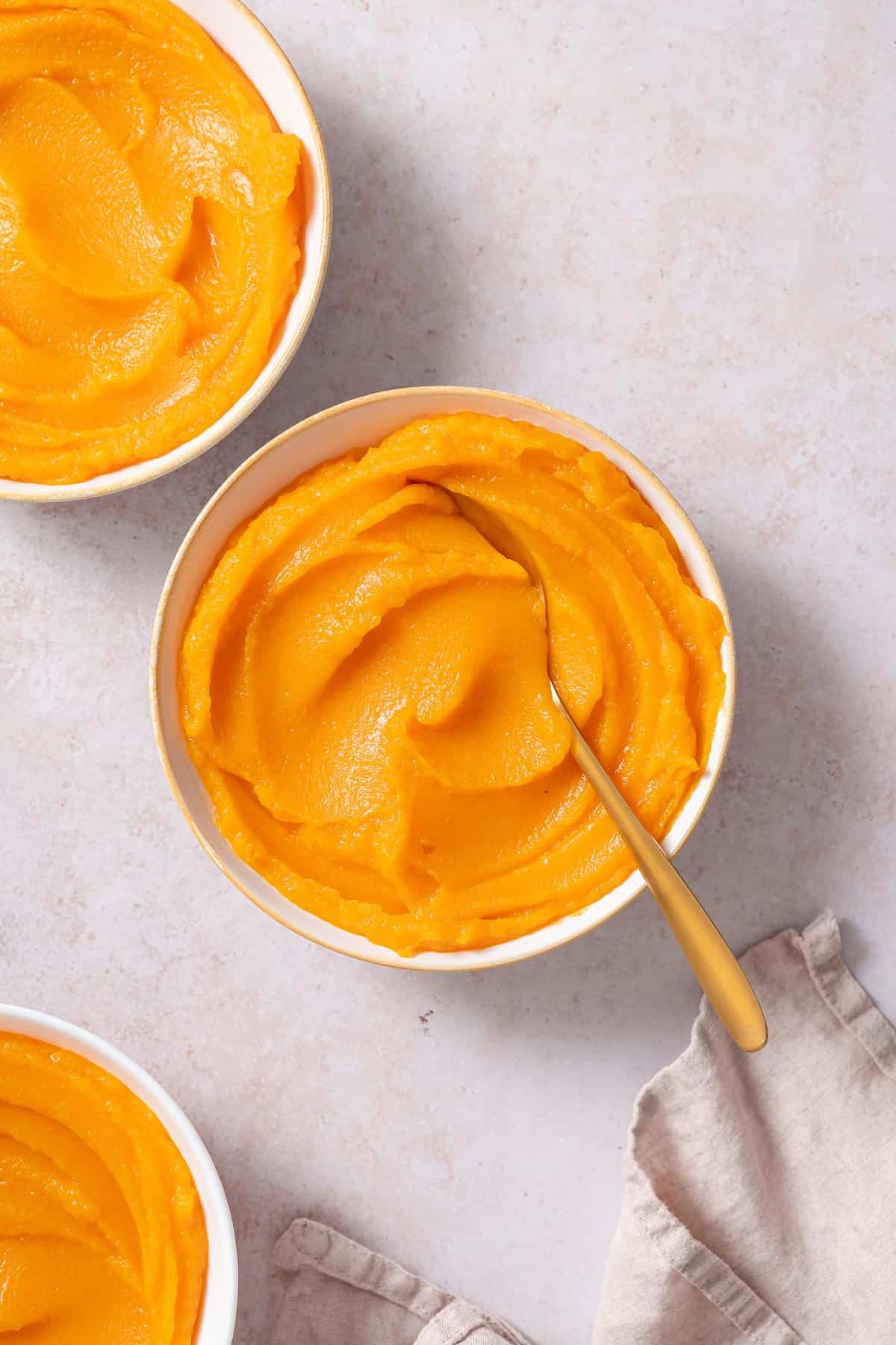 A bowl of homemade pumpkin puree with a spoon inside with two more bowls off to the side.