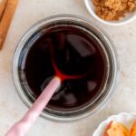 Overhead view of a jar of apple brown sugar syrup with sliced apples, brown sugar, and cinnamon sticks to the side.