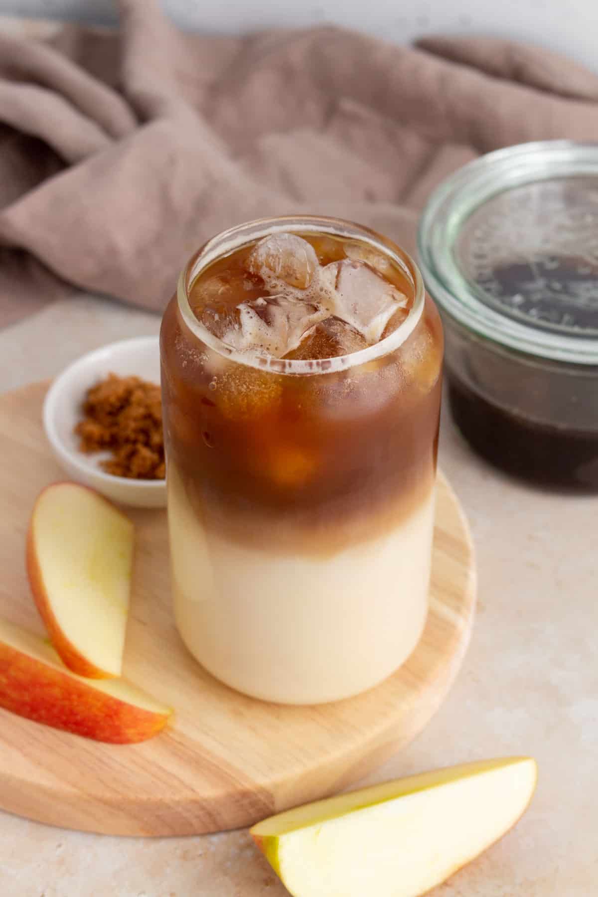 A slightly overhead view of a glass of iced apple crisp oatmilk macchiato with apple slices and syrup off to the side.