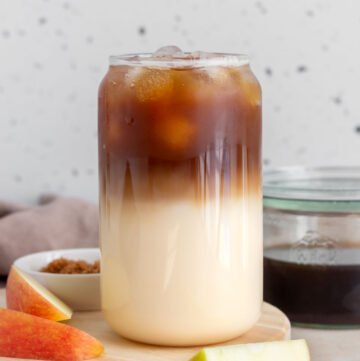 A glass of iced apple crisp oatmilk macchiato showing the two layers with some sliced apples in the front.