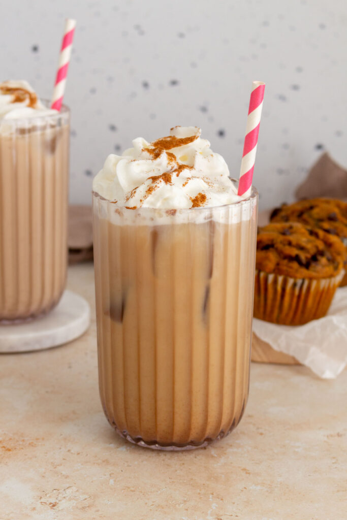 A glass of iced pumpkin spice latte with whipped cream and pumpkin pie spice sprinkled on top with a second glass and pumpkin muffins in the background.