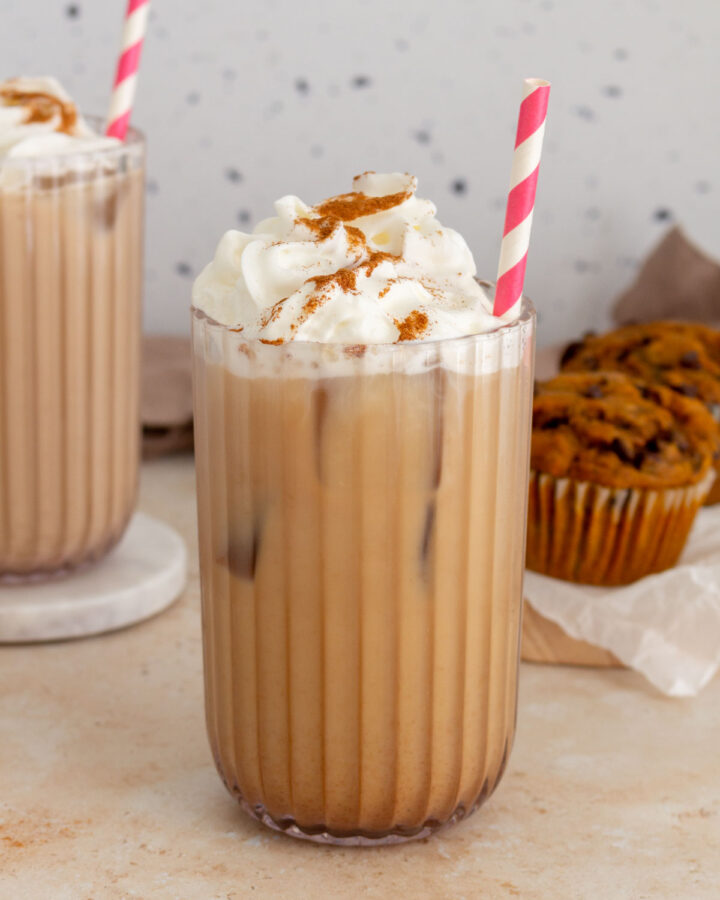A glass of iced pumpkin spice latte with whipped cream and pumpkin pie spice sprinkled on top with a second glass and pumpkin muffins in the background.