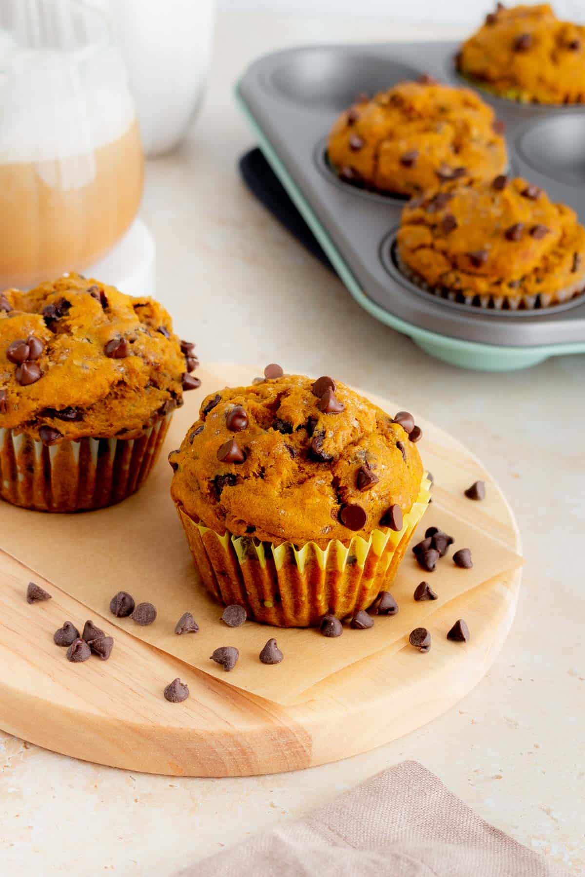 Two pumpkin banana muffins on a wooden board with three in a muffin tin in the background along with a mug of coffee.
