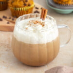 A mug of pumpkin spice latte with whipped cream and a sprinkle of pumpkin pie spice on top. A cinnamon stick inserted. Muffins in the background.