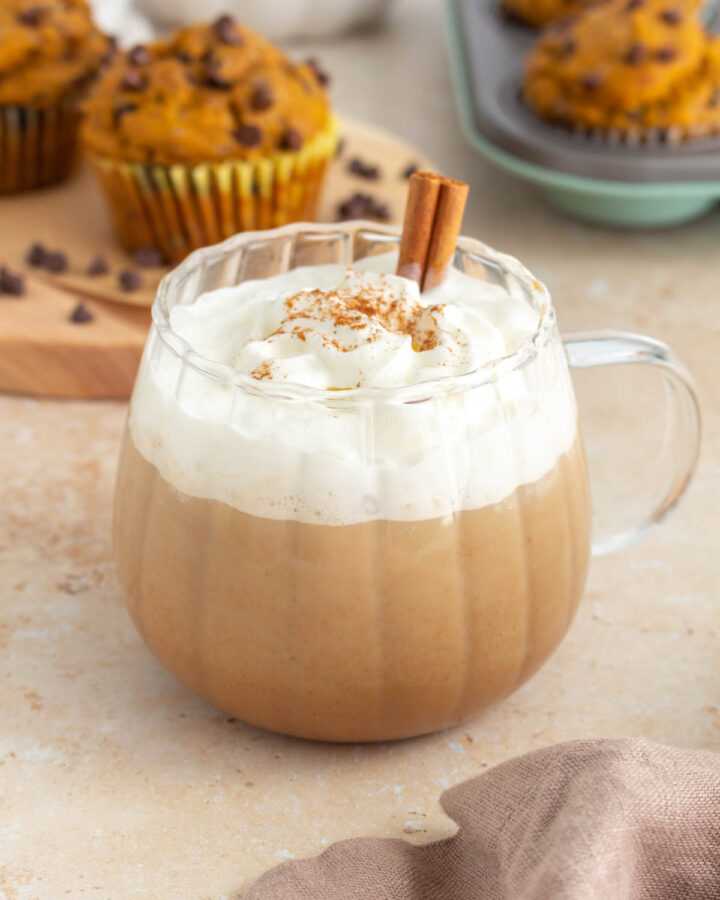 A mug of pumpkin spice latte with whipped cream and a sprinkle of pumpkin pie spice on top. A cinnamon stick inserted. Muffins in the background.