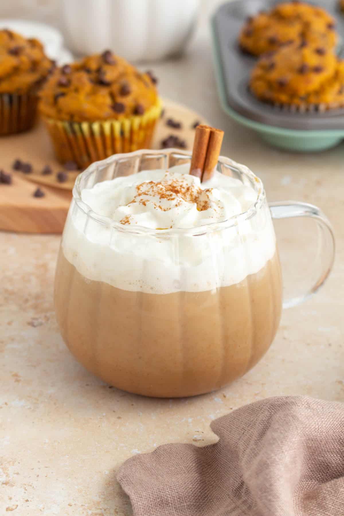 A mug of pumpkin spice latte with whipped cream and a sprinkle of pumpkin pie spice on top. A cinnamon stick inserted. Muffins in the background.