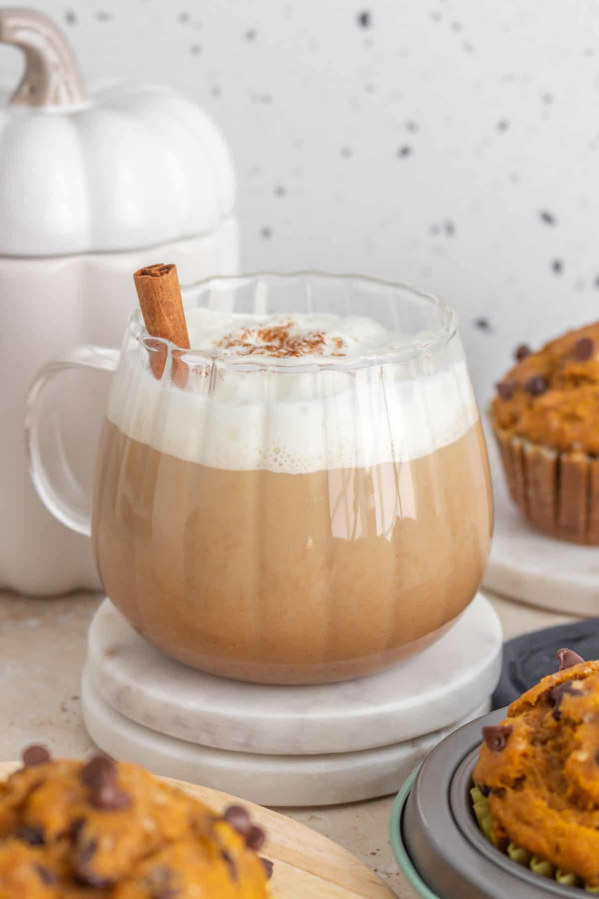 A mug of pumpkin spice latte with a cinnamon stick inserted, surrounded by muffins.
