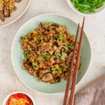 A plate of bulgogi fried rice with a pair of chopsticks on the plate. Green onions and kimchi on the side.