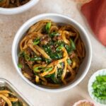 A bowl of black pepper udon with beef topped with sesame seeds and green onions surrounded by garnishes in bowls and a drink.