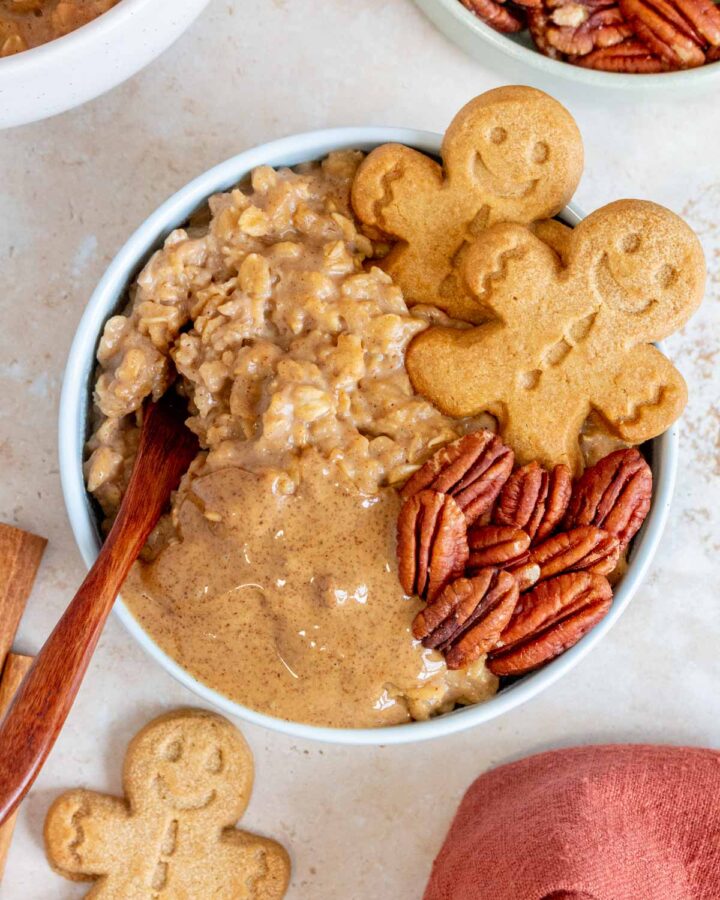 Overhead view of a bowl of gingerbread oatmeal topped with gingerbread cookies, pecans, and almond butter with a spoon inserted.