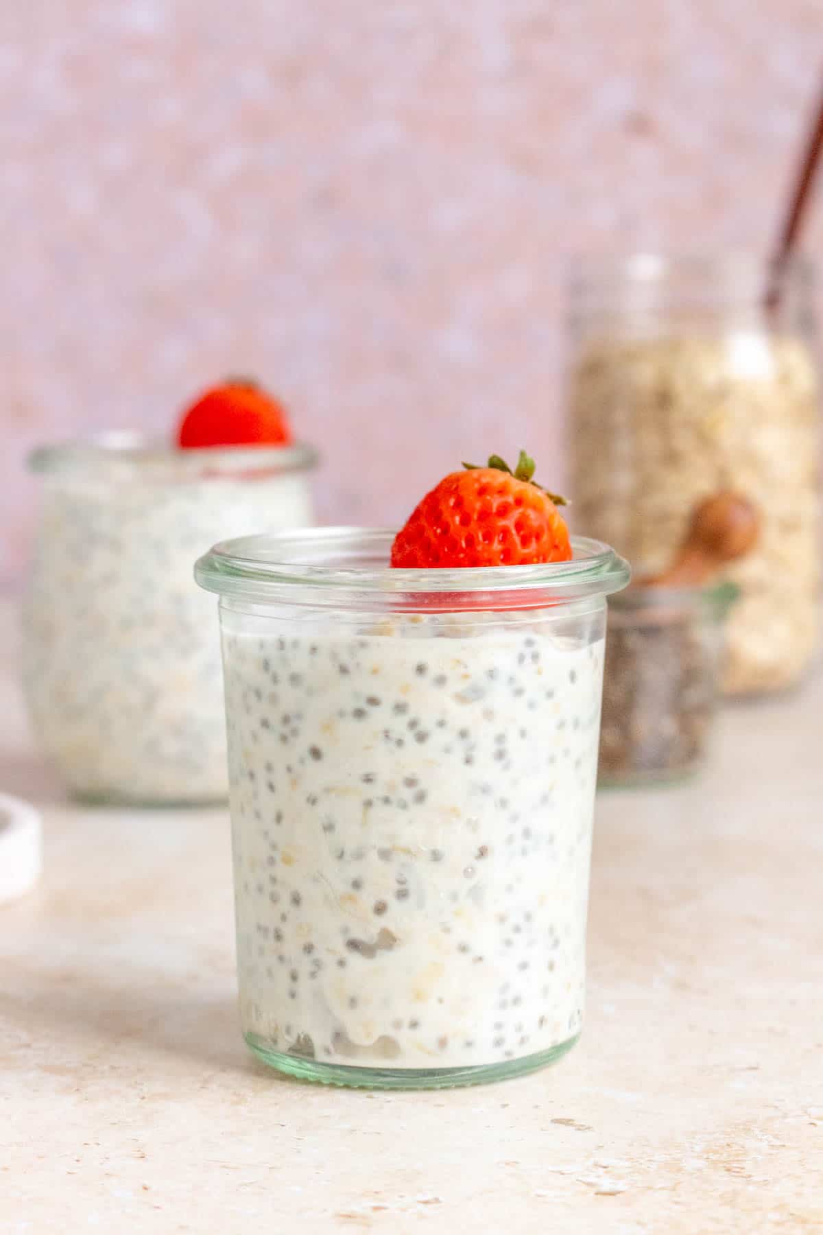 A jar of kefir overnight oats with a strawberry on top with another jar in the background out of focus along with a jar of chia seeds and rolled oats.