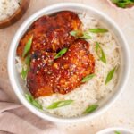 Overhead view of a bowl of rice with two tangy honey garlic chicken thighs topped with green onions. Rice, green onions, spinach, and sesame seeds off to the side.