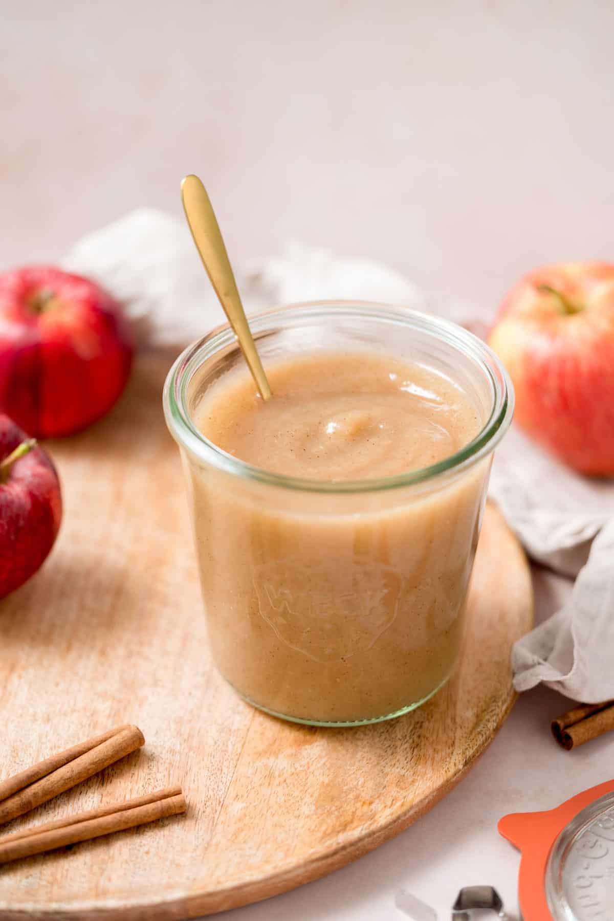 A jar of unsweetened applesauce with a spoon inside surrounded by apples.