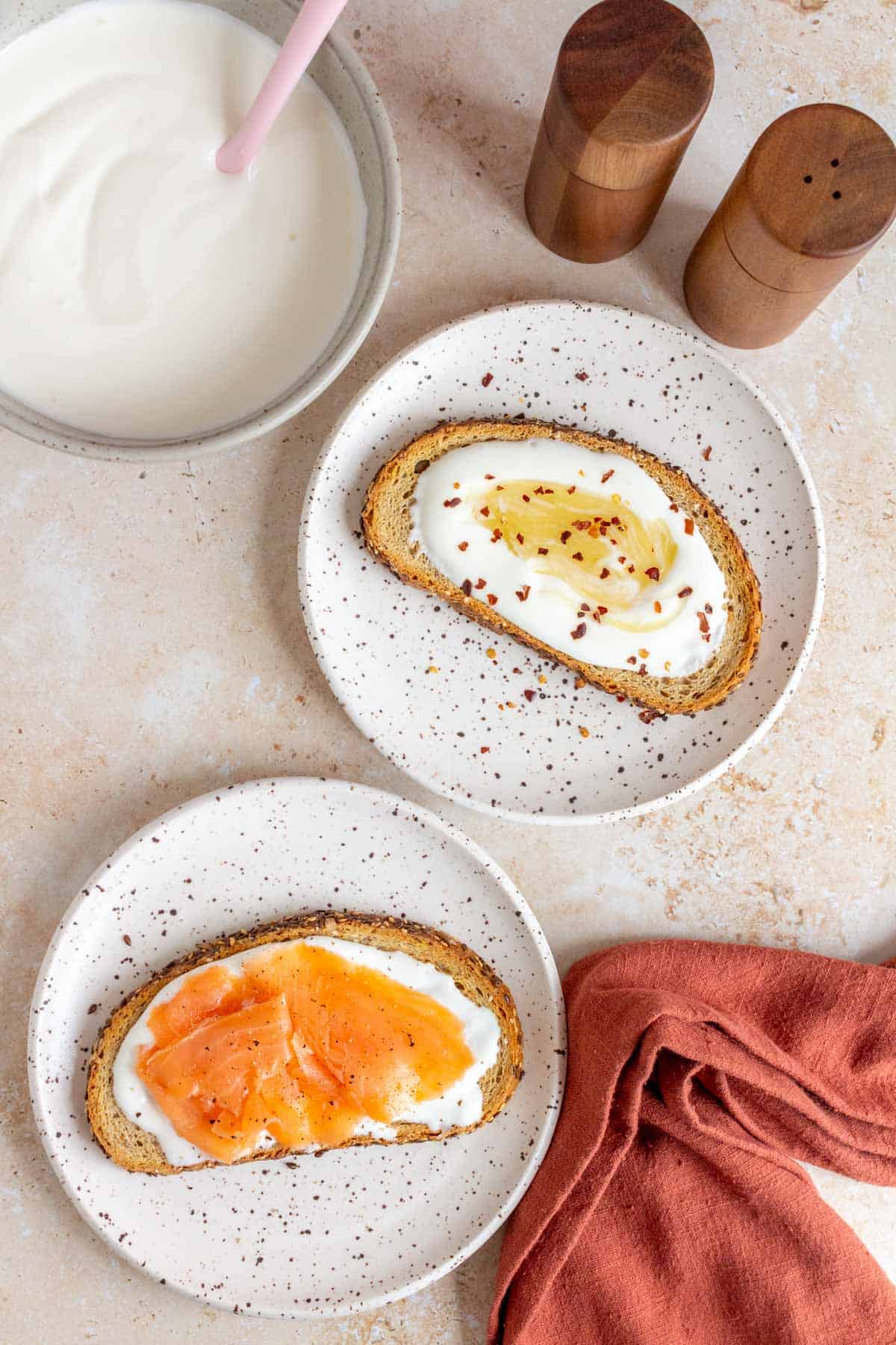 A plate with a piece of toast with whipped cottage cheese, honey, and red chili flakes beside another plate of toast with whipped cottage cheese topped with smoked salmon and pepper.
