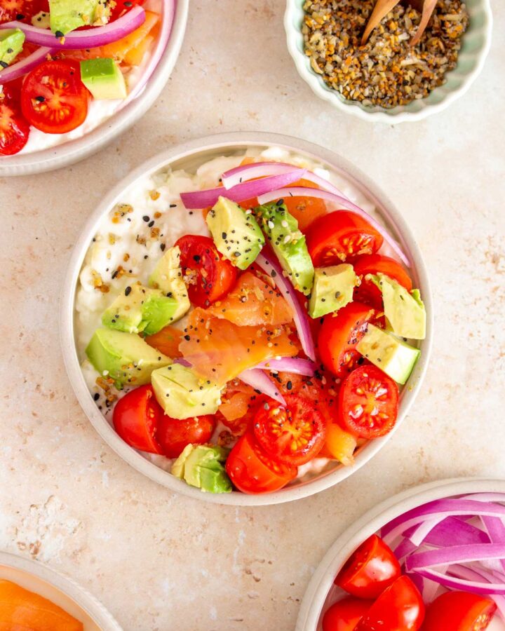 Overhead view of a bowl of cottage cheese and salmon surrounded by another plate and a plate of toppings along with a small bowl of everything but the bagel seasoning.