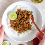 A plate of rice with ginger honey pork over top along with a lime. A hand holding a spoon tucked into the plate.