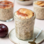 A jar of plum overnight oats topped with plum puree and sliced plums on a small plate with a spoon beside it.