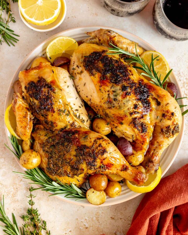 A platter with a roasted half chicken over potatoes, rosemary, and lemon slices.