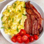 A plate of scrambled eggs with cottage cheese topped with green onions along with some strips of crispy bacon and cut grape tomatoes.