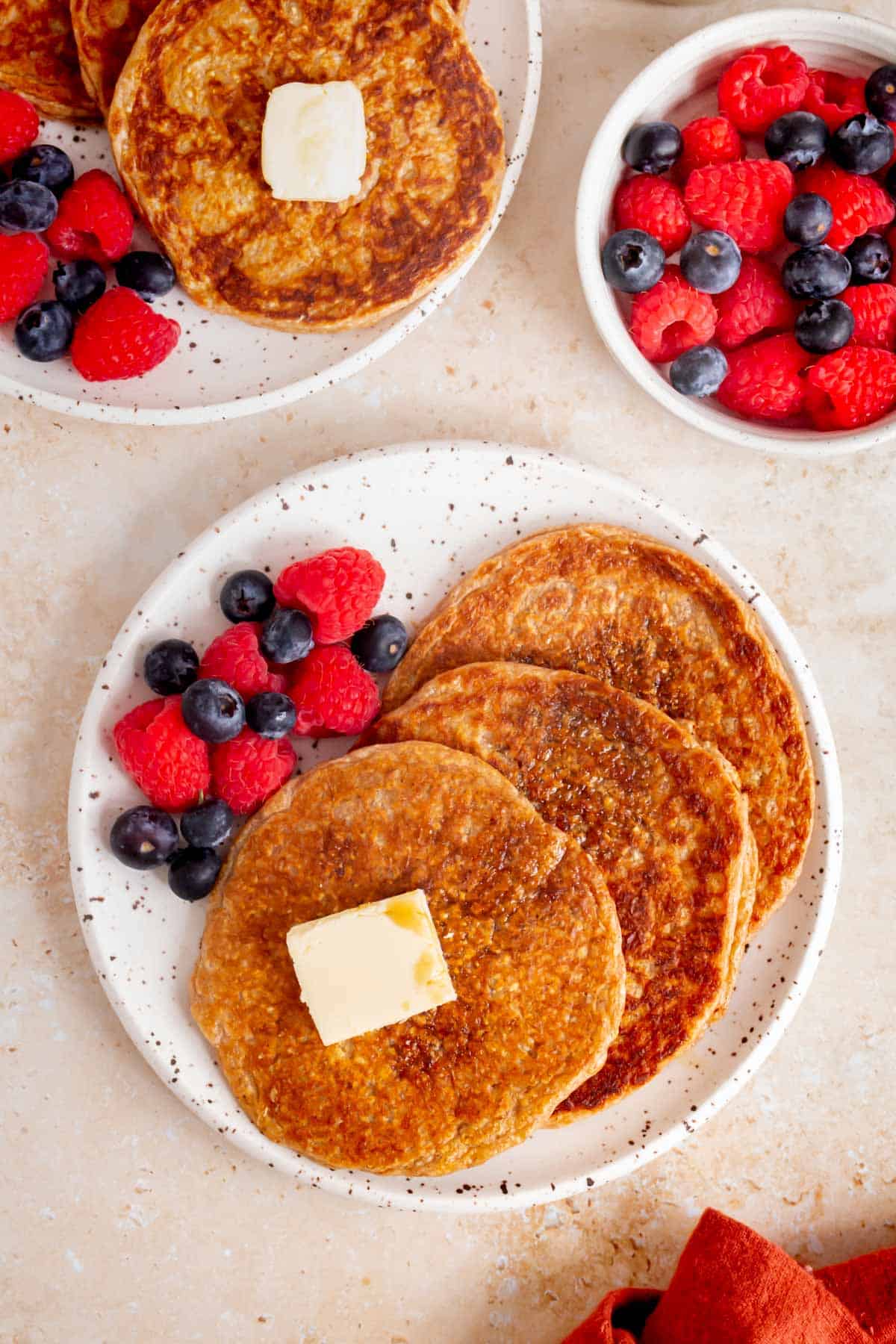 Overhead view of two plates of cottage cheese oatmeal pancakes with a bowl of berries on the side.