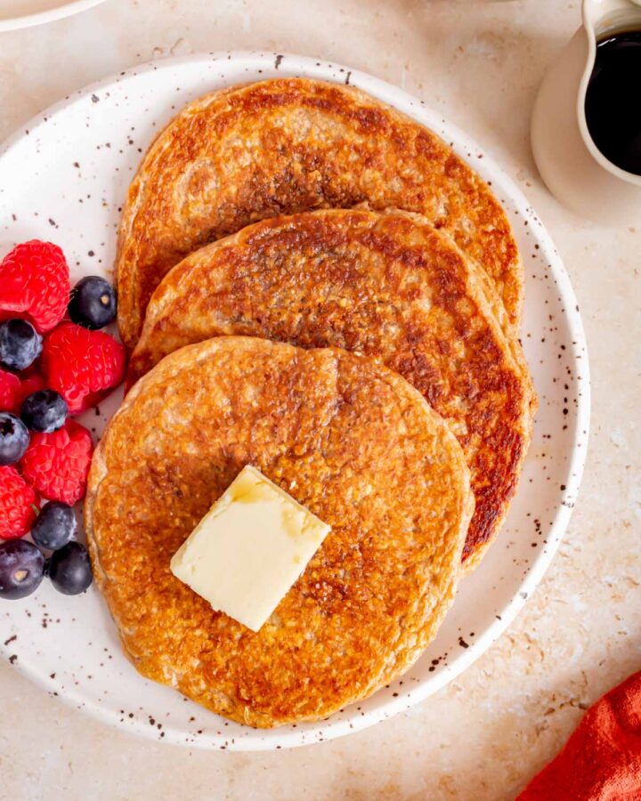 A close view of three cottage cheese oatmeal pancakes on a plate with syrup and a pat of butter with berries beside them.