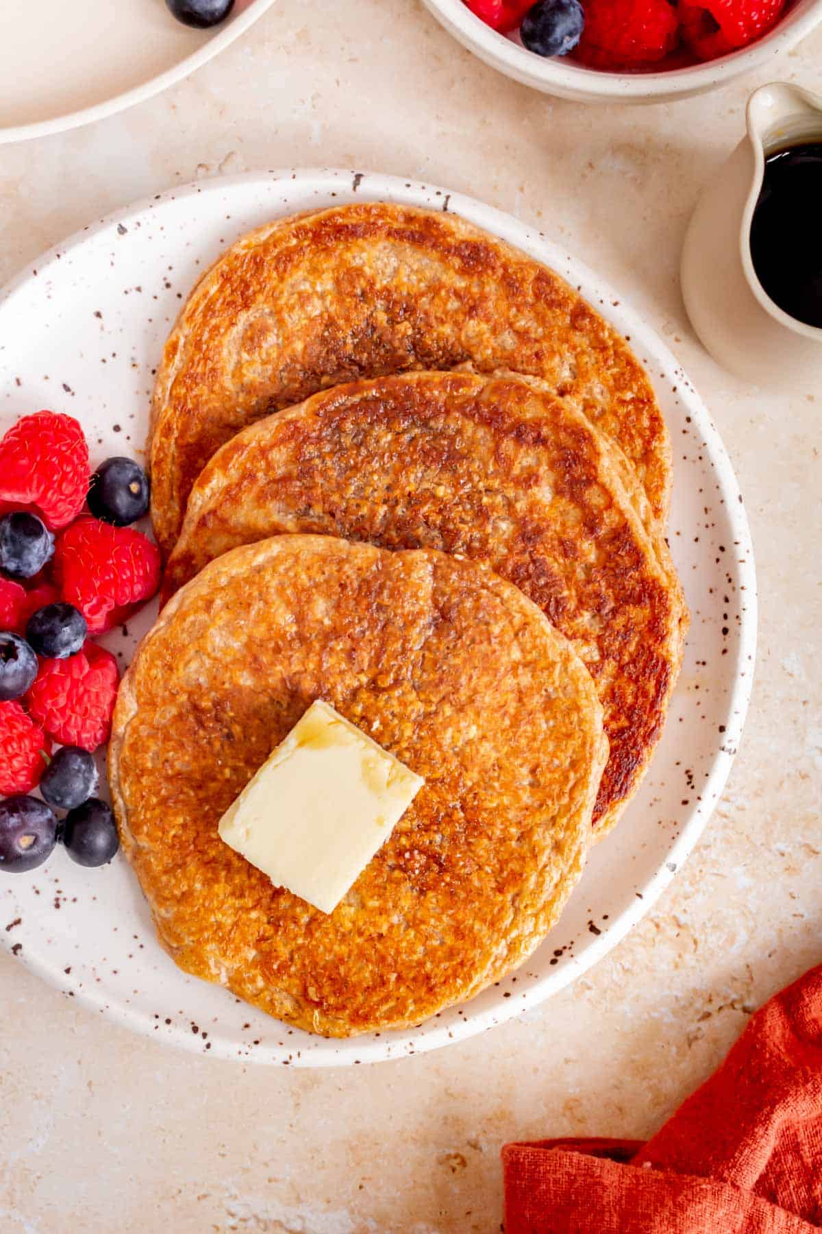 A close view of three cottage cheese oatmeal pancakes on a plate with syrup and a pat of butter with berries beside them.