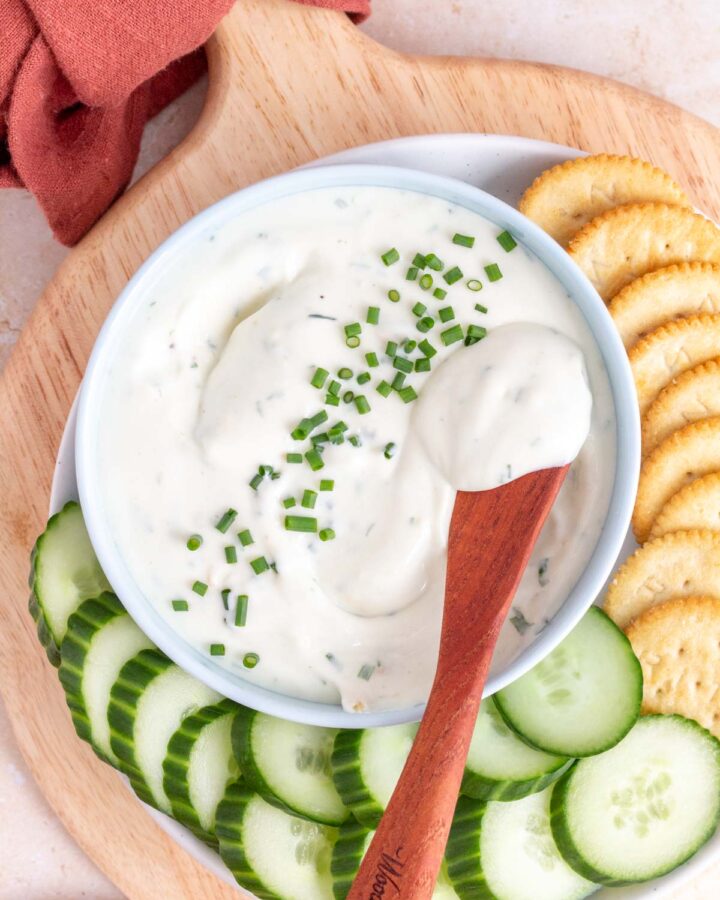 Overhead view of a bowl of cottage cheese with chives and roasted garlic with a spreader on top. Cucumbers and crackers on the side.
