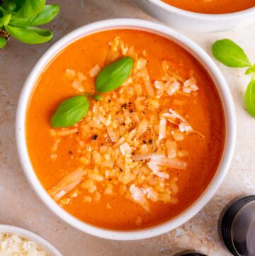 Overhead view of a bowl of high protein tomato soup topped with grated parmesan cheese, freshly cracked pepper, and basil leaves.