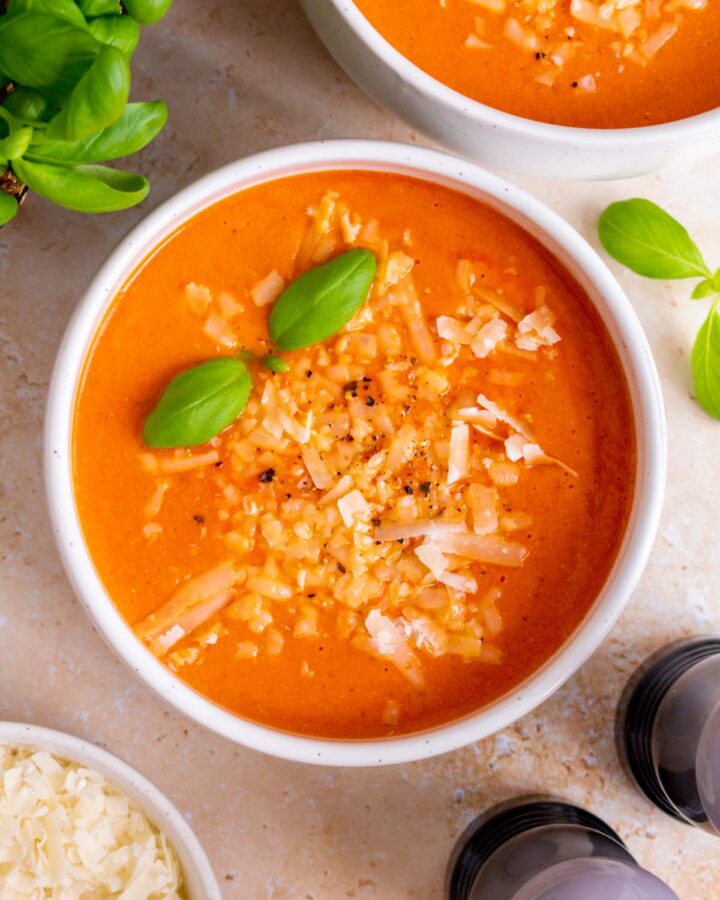 Overhead view of a bowl of high protein tomato soup topped with grated parmesan cheese, freshly cracked pepper, and basil leaves.