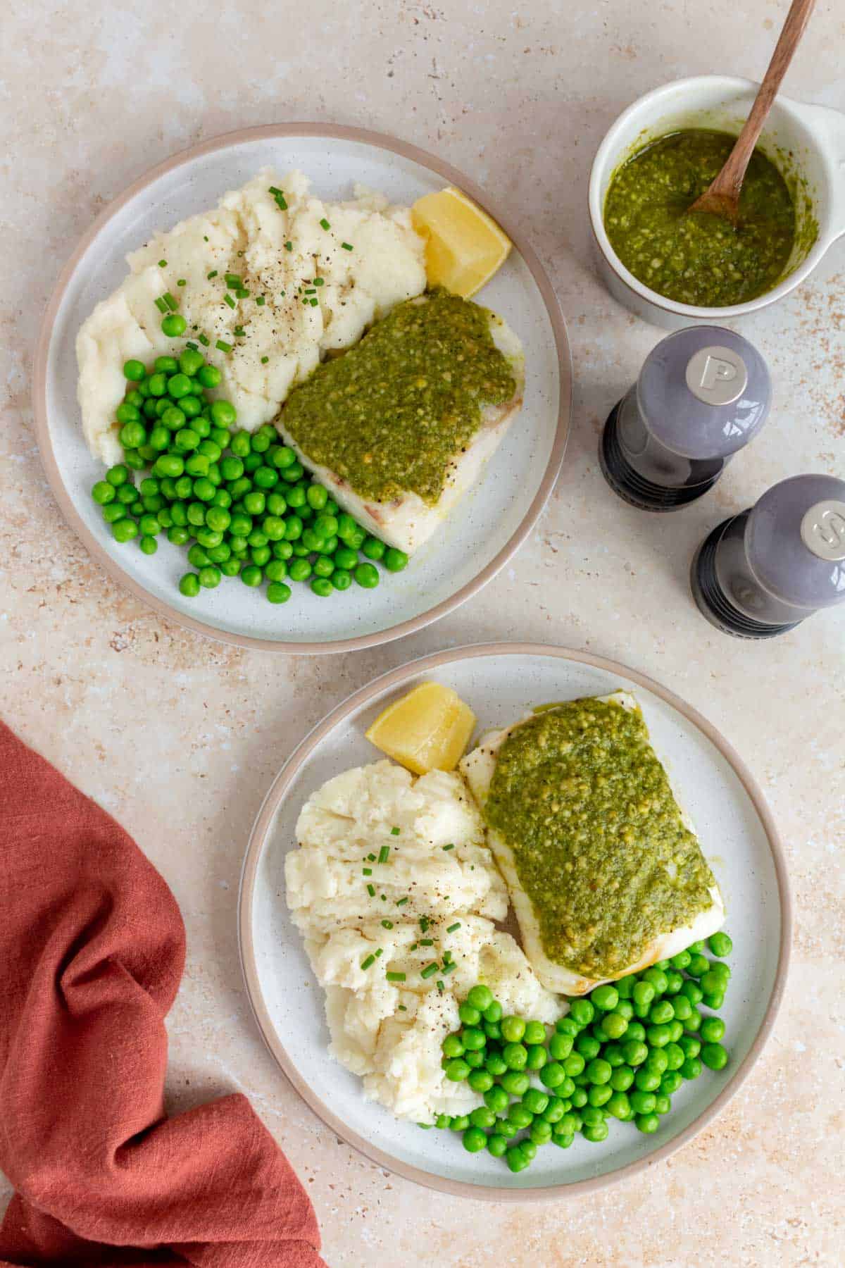 Two plates of pesto cod with mashed potatoes, peas, and lemon wedges. A bowl of pesto and salt and pepper grinder off to the side.