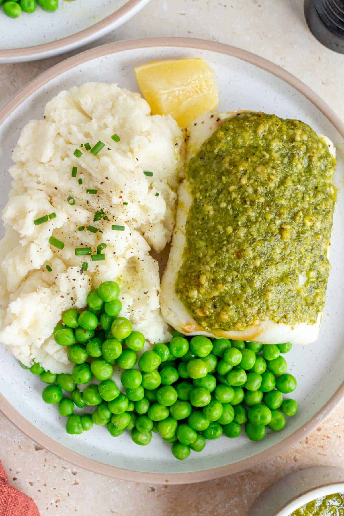 A plate with mashed potatoes, peas, pesto cod, and a lemon wedge.