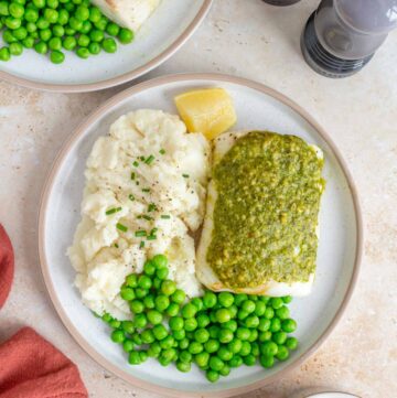 Overhead view of a plate with pesto cod, mashed potatoes, peas, and a lemon wedge. Second plate off to the side. Salt and pepper shakes to the side with a bowl of pesto.