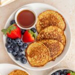 A plate with 4 pancakes along with maple syrup and berries. A bowl of berries off to the side.