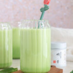 A glass cup of matcha protein shake on a coaster with a floral stir stick. Matcha tin in the background and spinach scattered.